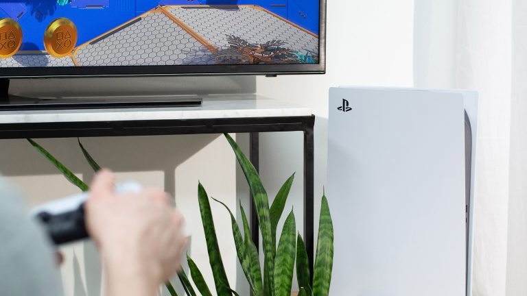 Eine Person sitzt vor einem Fernseher und spielt etwas auf einer PlayStation 5, die im Fokus der Kamera ist.
