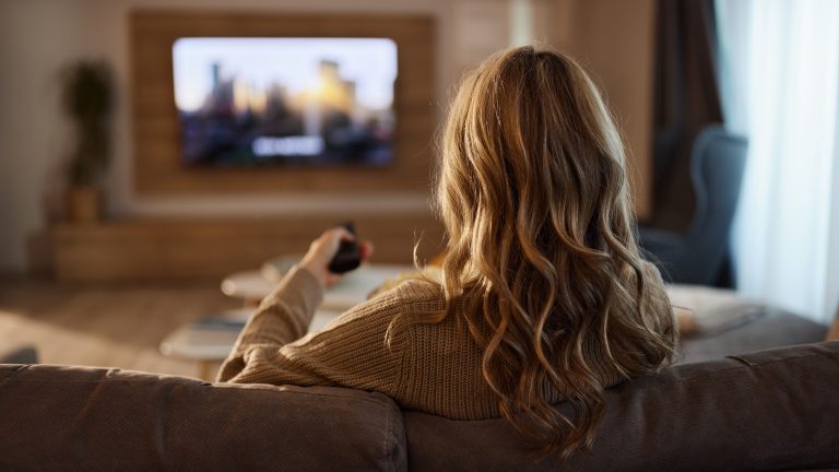 TVs von Hisense, Sender suchen und sortieren Eine Person sitzt mit einer Fernbedienung in der Hand auf einem Sofa und schaut fern.