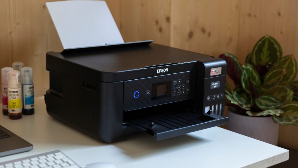 Ein schwarzer Epson EcoTank-Drucker steht auf Schreibtisch, daneben Tintenflaschen, mit Pflanzen im Hintergrund.
