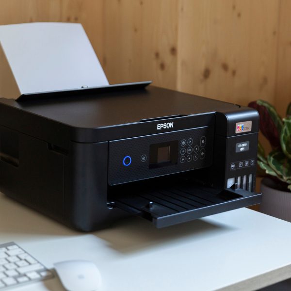 Ein schwarzer Epson EcoTank-Drucker steht auf Schreibtisch, daneben Tintenflaschen, mit Pflanzen im Hintergrund.