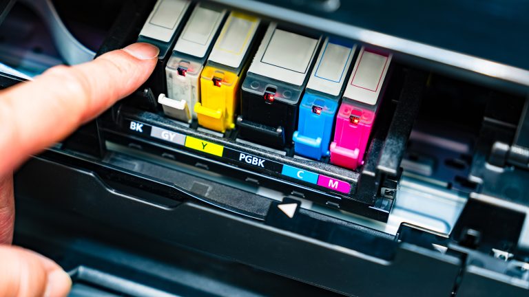 Tintenstrahldrucker, Patrone oder Tank - so triffst du die richtige Wahl Eine Person zeigt auf die farbigen Einzelpatronen (Schwarz, Gelb, Cyan, Magenta) eines Tintenstrahldruckers im geöffneten Patronenfach.