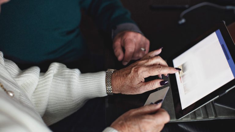 Zwei Personen bedienen gemeinsam ein Tablet und zeigen auf den Bildschirm; Nahaufnahme von H&auml;nden und Ger&auml;t auf einem Tisch.