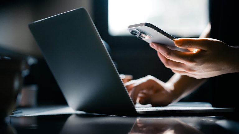 Nahaufnahme einer Hand, die ein Smartphone h&auml;lt. Die andere Hand bedient parallel dazu einen Laptop.