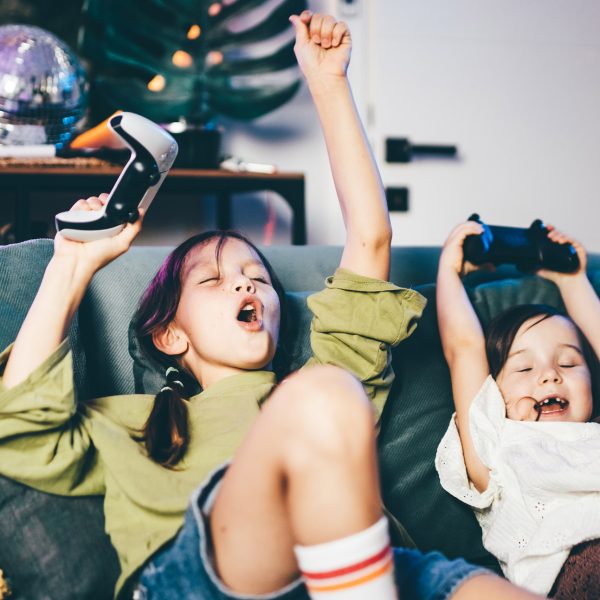 Zwei Kinder mit Dualsense-Controllern sitzen auf einem Sofa und rei&szlig;en die Arme freudig in die Luft.