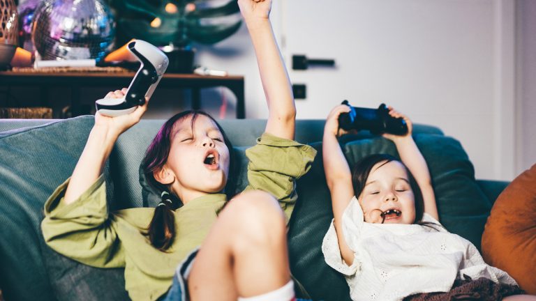 Zwei Kinder mit Dualsense-Controllern sitzen auf einem Sofa und rei&szlig;en die Arme freudig in die Luft.