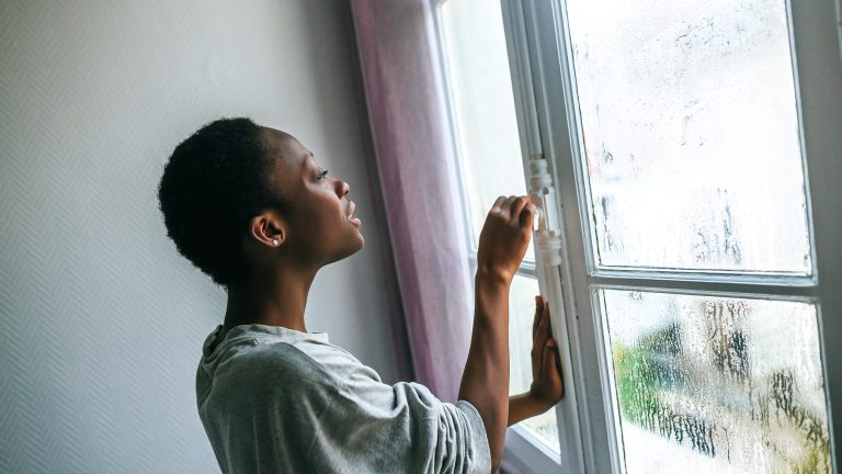 Eine Person steht vor einem Fenster und blickt nach drau&szlig;en. Die Hand liegt auf dem Fenstergriff als wolle sie dieses &ouml;ffnen.