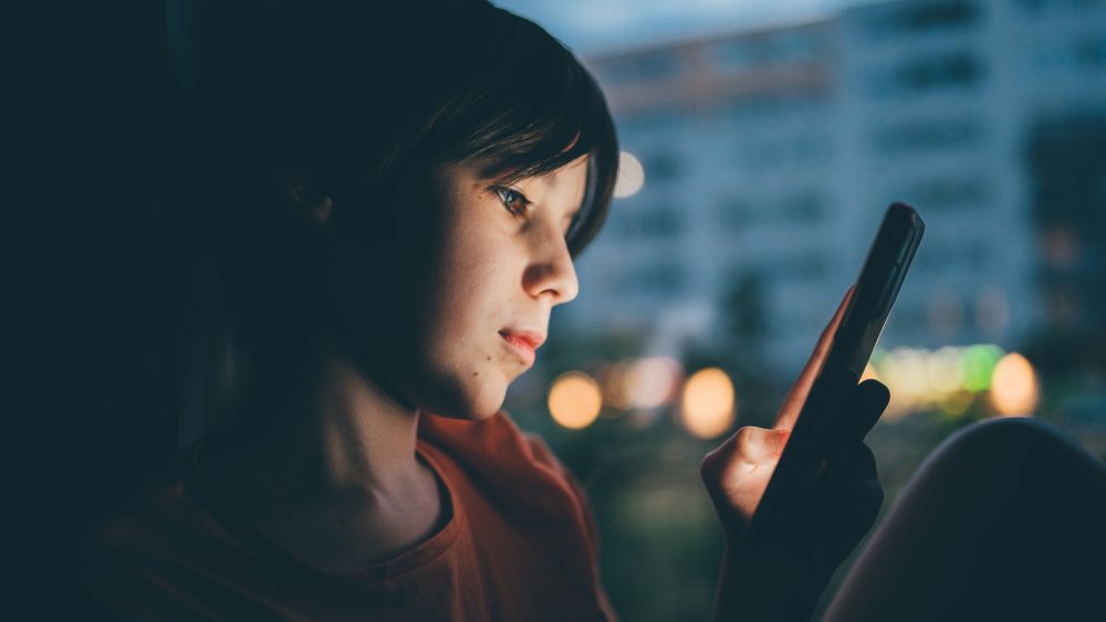 Eine Person sitzt in einem dunklen Raum und blickt auf ein Smartphone.