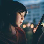 Eine Person sitzt in einem dunklen Raum und blickt auf ein Smartphone.