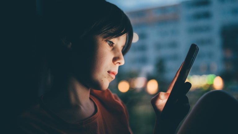 Eine Person sitzt in einem dunklen Raum und blickt auf ein Smartphone.