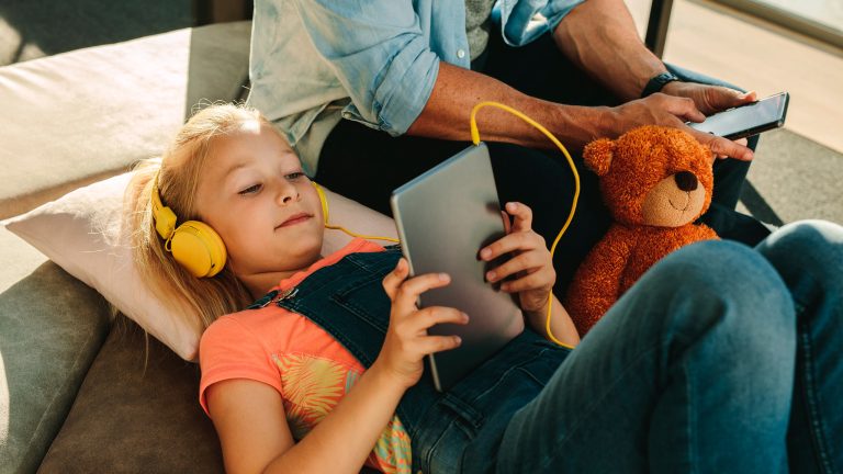 Eine Person mit Kopfhörern liegt auf einem Sofa und hält ein Tablet. Daneben sitzt eine weitere Person mit einem Smartphone; zwischen beiden liegt ein Plüschtier.