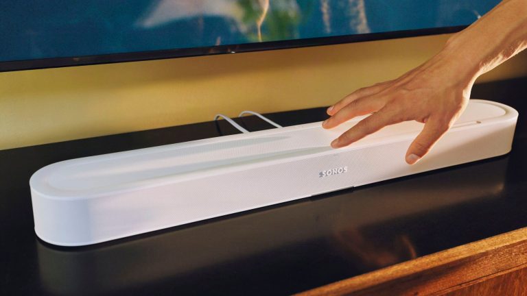 Eine Sonos Beam Ultra in Wei&szlig; steht unter einem Fernseher. Eine Person bedient die Soundbar mit der Hand.