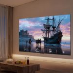 In einem Wohnzimmer ist ein Fernseher an der Wand montiert, auf dem Bildschirm ist ein altes Segelschiff zu sehen.