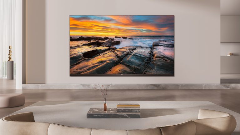 TCL-TV, Sendersuchlauf durchführen und Sender sortieren In einem Wohnzimmer hängt ein Fernseher an der Wand, auf dem Bildschirm sind felsige Küstenplatten zu sehen, über die Wellen hinwegspülen, während der Himmel in warmen Farben eines Sonnenuntergangs leuchtet.