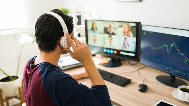 Eine Person mit Headset sitzt am Schreibtisch vor einem Monitor auf dem eine Videokonferenz zu sehen ist.