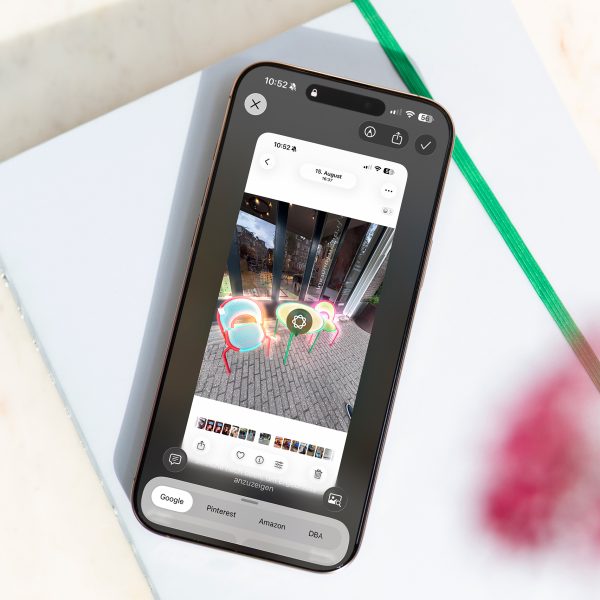 Ein iPhone liegt auf einem wei&szlig;en Notizbuch. Auf dem Bildschirm ist ein Screenshot von Outdoor-M&ouml;beln zu sehen. Sie sind markiert f&uuml;r eine visuelle Suche.
