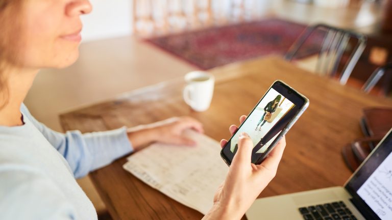 Eine Person sitzt an einem Tisch. In ihrer Hand h&auml;lt sie ein Smartphone, auf dem das Bild ihrer Video-Doorbell eine Person zeigt, die vor ihrer Haust&uuml;r steht.