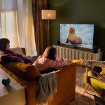 Eine Person sitzt mit zwei Kindern vor einem Fernseher, auf dem eine Naturdoku läuft. Unter dem Fernseher ist ein Apple TV 4K zu sehen, daneben zwei HomePods.