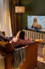 Eine Person sitzt mit zwei Kindern vor einem Fernseher, auf dem eine Naturdoku läuft. Unter dem Fernseher ist ein Apple TV 4K zu sehen, daneben zwei HomePods.