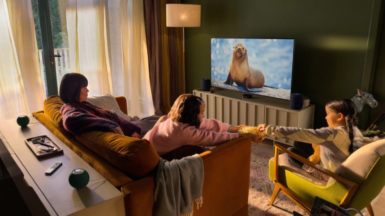 Eine Person sitzt mit zwei Kindern vor einem Fernseher, auf dem eine Naturdoku läuft. Unter dem Fernseher ist ein Apple TV 4K zu sehen, daneben zwei HomePods.