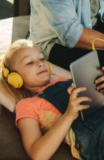 Kopfhörer für Kinder, 4 Modelle für mobilen Hörspaß Ein Kind hört über Kopfhörer etwas von einem Tablet. Daneben sitzt ein Elternteil.