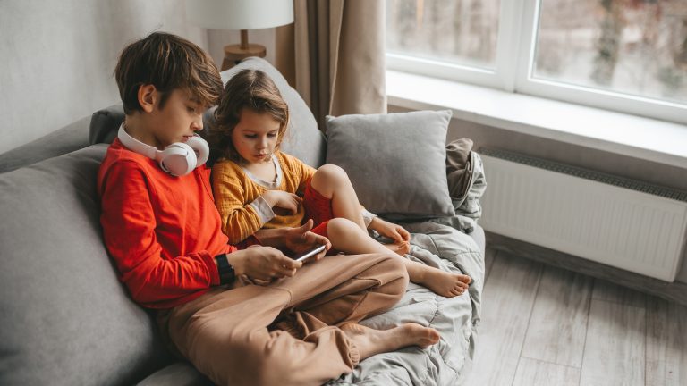Zwei Kinder sitzen auf einem Sofa und schauen zusammen auf ein Smartphone. Eines der Kinder trägt Kopfhörer.