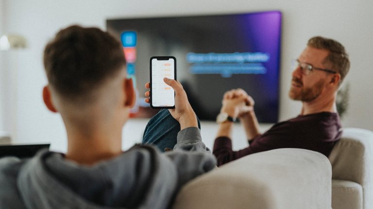 Smart-Home-Security, So schützt du dich vor Hackern Eine Person mit dem Smartphone in der Hand sitzt auf einem Sofa. Vor ihr sitzt eine weitere Person.