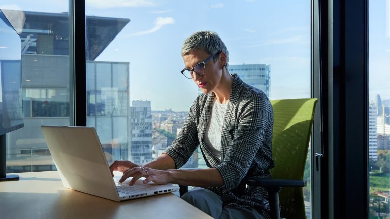 Eine Person sitzt an einem Tisch vor einem Notebook in einem Büro mit großen Fenstern.